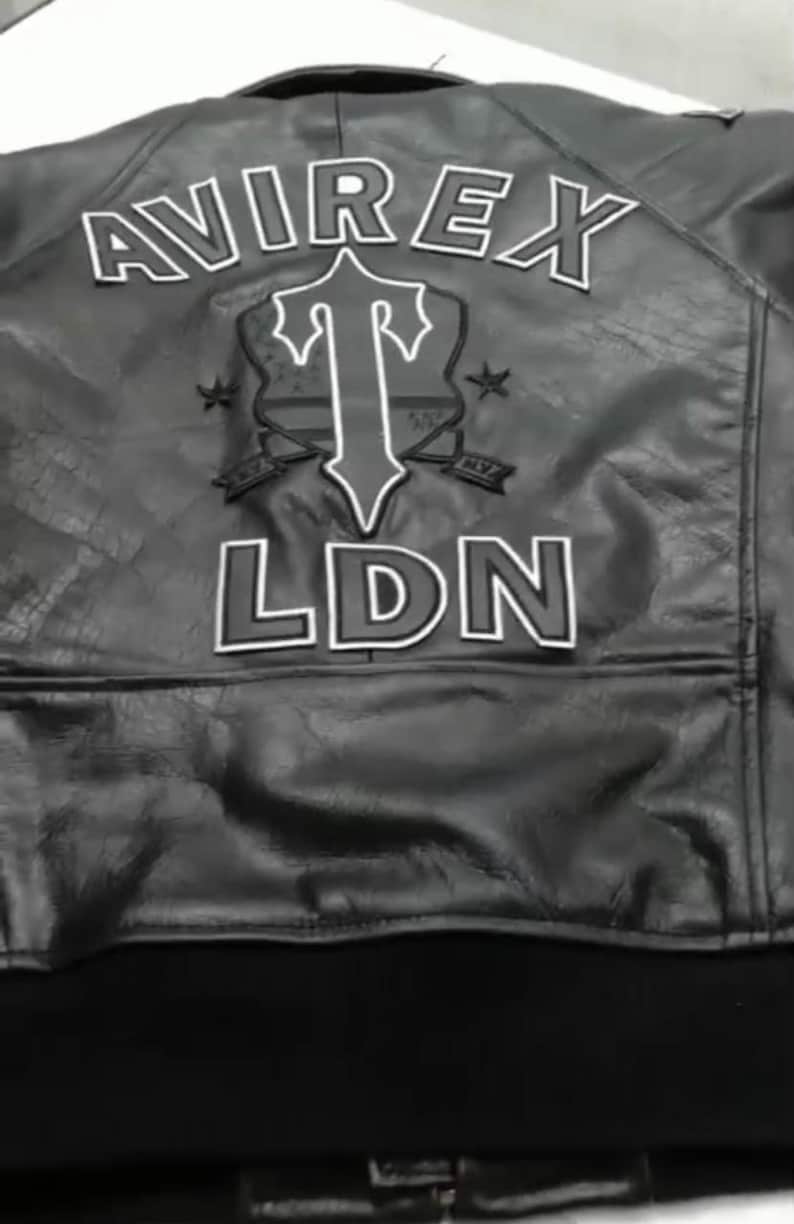 May include: Black leather jacket with white lettering and a shield design. The jacket features the brand name "AVIREX" across the top, with "LDN" at the bottom. A shield graphic is in the center.