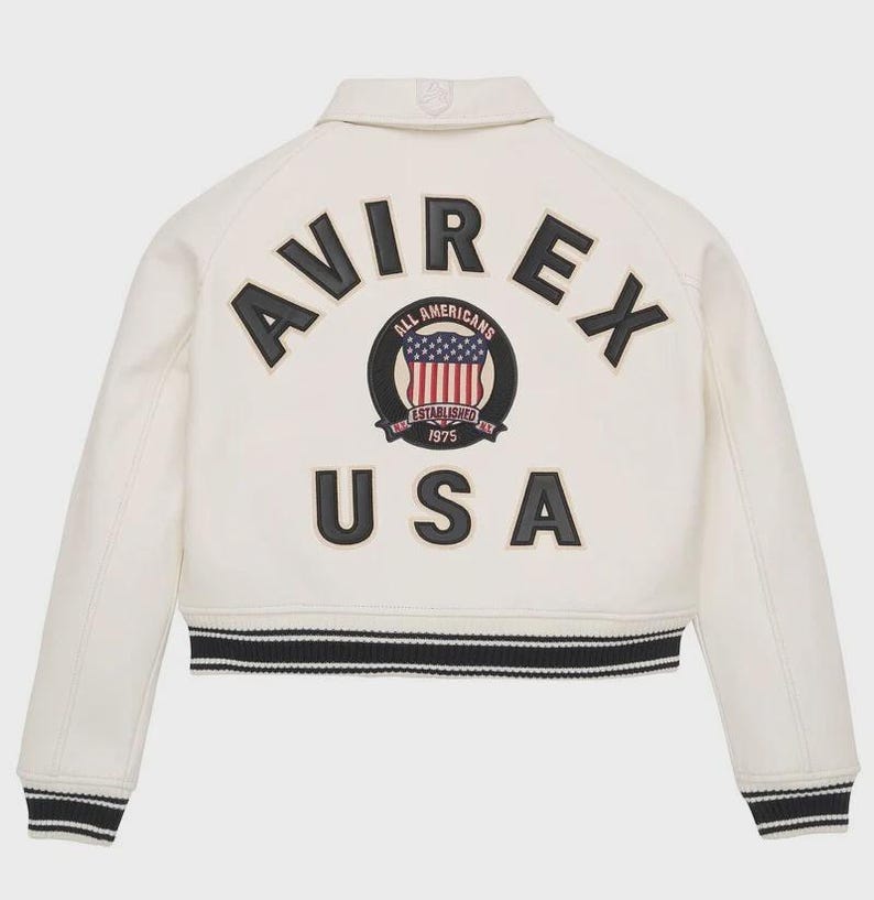 May include: Cream-colored jacket with black and white striped trim. The back features the word "AVIREX" in black letters above a circular emblem with a US flag design and the word "USA" in black letters.