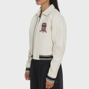 May include: Cream-colored bomber jacket with a zipper closure, collar, and black and white striped trim on the cuffs and hem. Features a patch on the chest with the text "AVIREX" and an American flag design.
