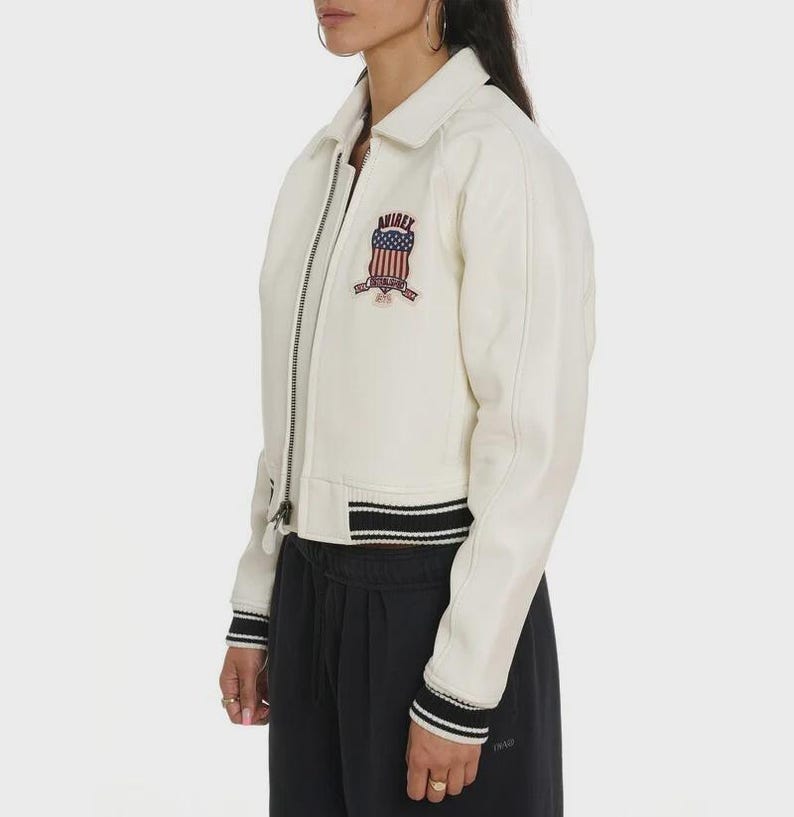 May include: Cream-colored bomber jacket with a zipper closure, collar, and black and white striped trim on the cuffs and hem. Features a patch on the chest with the text "AVIREX" and an American flag design.
