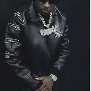 May include: Black leather jacket with white lettering on the sleeves and a patch on the chest. The jacket has a collar and a zipper closure. A silver chain necklace and a wristwatch are also visible.