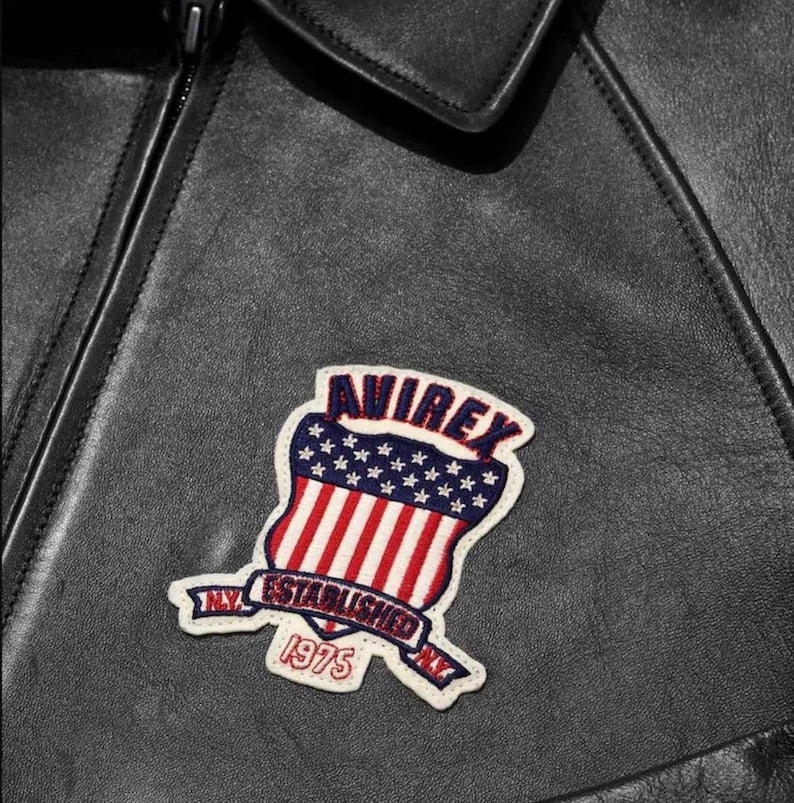 May include: Close-up of a black leather jacket with a patch. The patch is a shield shape with red and white stripes, blue stars, and the word "AVIREX". Below the shield is a banner that reads "NY ESTABLISHED 1975".