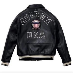May include: Black leather jacket with white lettering that reads "AVIREX" across the shoulders and "USA" below. A circular patch with the words "ALL AMERICANS ESTABLISHED 1975" and a US flag design is centered. The jacket has a ribbed collar and cuffs.
