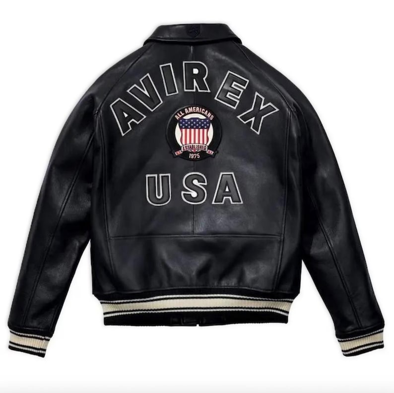 May include: Black leather jacket with white lettering that reads "AVIREX" across the shoulders and "USA" below. A circular patch with the words "ALL AMERICANS ESTABLISHED 1975" and a US flag design is centered. The jacket has a ribbed collar and cuffs.