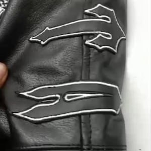 May include: Black leather jacket sleeve with white decorative stitching. The jacket features several stylized flame-shaped designs outlined in white. The leather appears to be smooth and the stitching is precise.