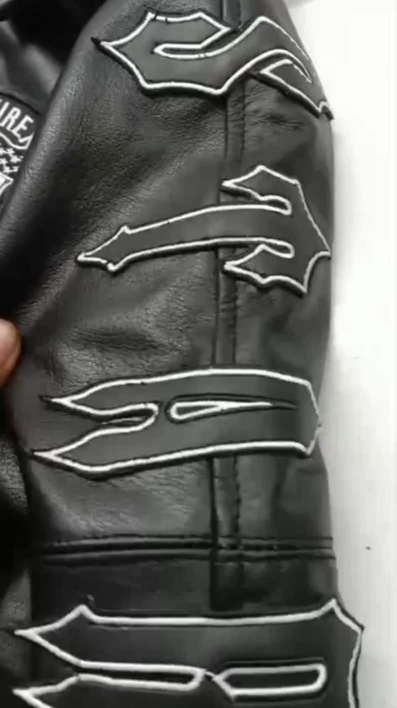 May include: Black leather jacket sleeve with white decorative stitching. The jacket features several stylized flame-shaped designs outlined in white. The leather appears to be smooth and the stitching is precise.
