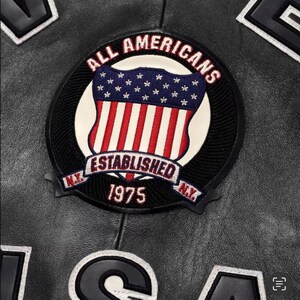May include: Close-up of a black leather jacket featuring a circular patch with the text "ALL AMERICANS" and "ESTABLISHED 1975." The patch displays a shield design with the American flag. The jacket also has other patches.