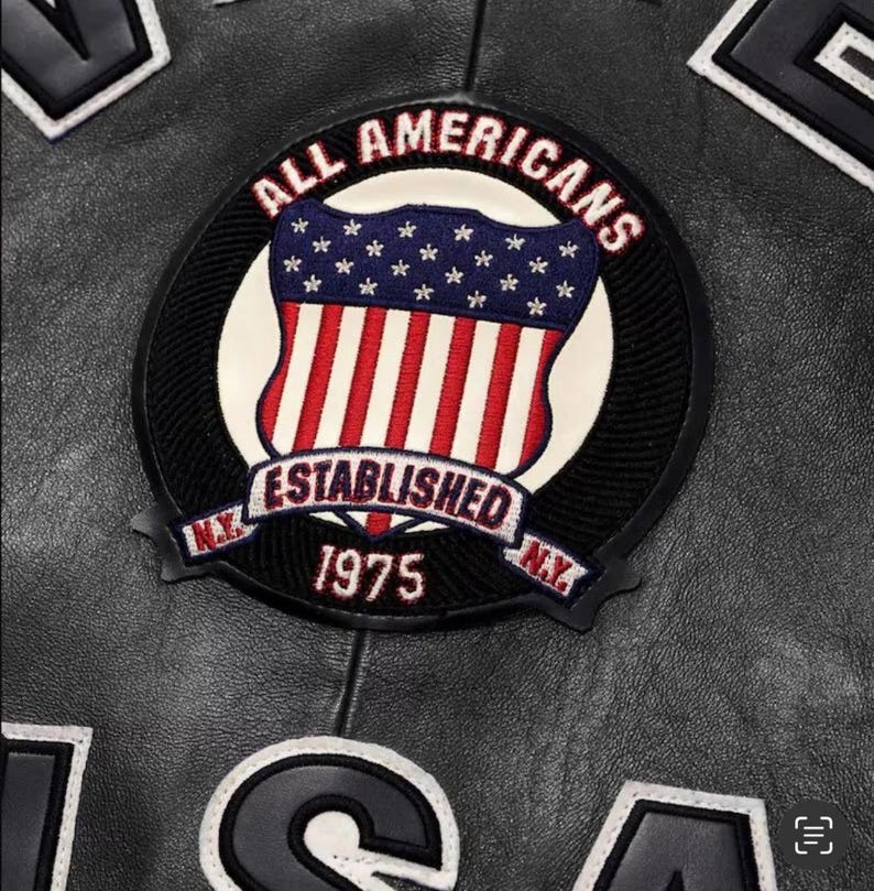 May include: Close-up of a black leather jacket featuring a circular patch with the text "ALL AMERICANS" and "ESTABLISHED 1975." The patch displays a shield design with the American flag. The jacket also has other patches.