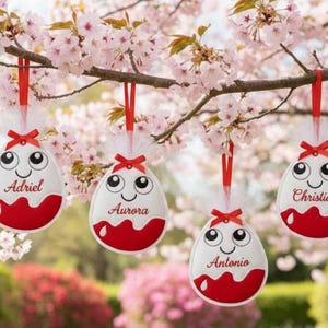 Könnte beinhalten: Vier hängende Ostereier-Dekorationen mit den Namen Adriel, Aurora, Antonio und Christian. Jedes Ei ist weiß mit rotem Boden, einem lächelnden Gesicht und einem roten Band. Sie hängen an einem blühenden Ast.
