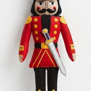May include: A felt nutcracker doll dressed in a red uniform with yellow buttons, black trousers, and a black hat with a yellow brim. The doll features a black mustache, red cheeks, and a silver sword tucked into a black belt across the chest.