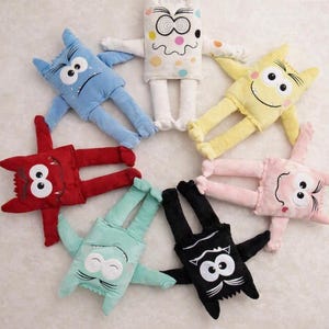 May include: A circular arrangement of seven plush monster toys in various colors including blue, white with polka dots, yellow, pink, black, light green, and red. Each soft toy features two large white eyes, stitched facial expressions, and limbs.