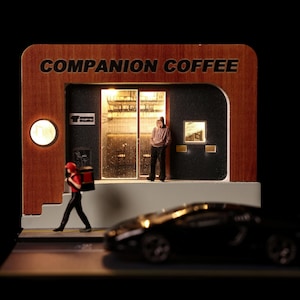 Può includere: Un diorama in miniatura di una caffetteria chiamata COMPANION COFFEE con facciata in legno, interni illuminati e porta a vetri. Una piccola figura con una camicia rossa passa davanti, mentre un'altra figura si trova sulla soglia. Un'auto giocattolo nera è in primo piano.