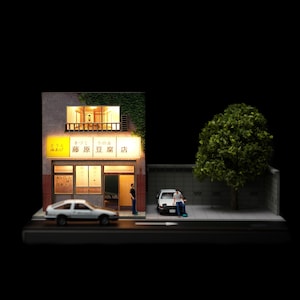 Peut inclure: Un diorama miniature d'un bâtiment à deux étages avec une enseigne jaune lumineuse portant l'inscription Fujiwara Tofu Shop en caractères japonais. La scène comprend deux petites figurines, une voiture blanche garée et un arbre vert.
