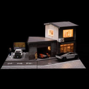 Puede incluir: Un diorama en miniatura que muestra un edificio de dos pisos con ventanas iluminadas, un garaje adjunto y una escena callejera. El modelo incluye un coche blanco y negro, un deportivo naranja, una camioneta plateada y dos pequeñas figuras humanas.
