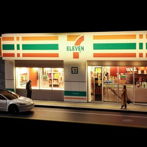 May include: A miniature scale model of a 7-Eleven convenience store featuring a white facade with orange and green horizontal stripes, illuminated glass storefront windows, a silver sedan parked in front, and two small figures on the sidewalk.
