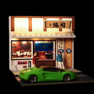 May include: A miniature diorama of a Japanese restaurant storefront with illuminated signage featuring the kanji for yakiniku. The scene includes two small figures standing outside the shop and a bright green sports car model in the foreground.
