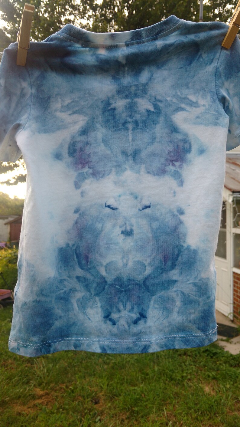 May include: A blue tie-dye t-shirt with a symmetrical pattern. The shirt is hanging on a clothesline.