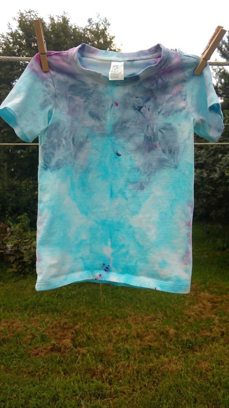 May include: A blue, purple, and pink tie-dye t-shirt hanging on a clothesline. The shirt has a light blue background with darker blue, purple, and pink swirls.