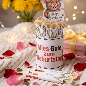 Puede incluir: Un juego de regalo de cumpleaños con un recipiente cilíndrico con la inscripción "Alles Gute zum Geburtstag" y un ratón de dibujos animados con un cartel de "Happy Birthday". Incluye dientes de león de papel, barritas de chocolate Kinder y pétalos de rosa.