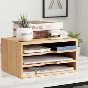 May include: A light-colored wooden desk organizer with three shelves. A small white pot with the word "CACTUSES" is on top, along with books and a stapler. The organizer is filled with papers and documents.