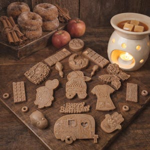 May include: A still life arrangement with cinnamon-sugar doughnuts, red apples, and brown wax melts in various shapes. A lit candle warmer is on the right. The wax melts include a camper, a hand, and a 'Happy Birthday' message.