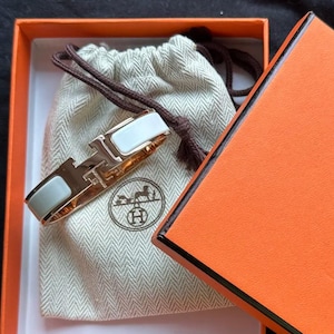 May include: A gold-tone bracelet with a white enamel inlay, presented in an orange box with the brand's logo. The bracelet is displayed inside a beige drawstring pouch. The box is open, revealing the bracelet and pouch.