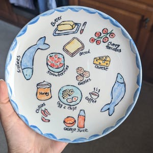 May include: A white ceramic plate with a blue scalloped edge, hand-painted with food illustrations. Includes sardines, butter, strawberries, bread, cookies, cheese, honey, chilies, egg & chips, coffee, and orange juice.