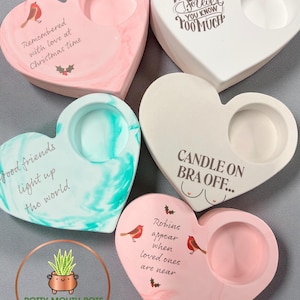 Könnte beinhalten: Herzförmige Kerzenhalter in verschiedenen Farben, darunter Rosa, Weiß und Türkis, mit Marmoreffekten. Jeder Halter hat eine kreisförmige Vertiefung für eine Kerze und dekorativen Text. Einige haben Vogel- und Stechpalmen-Illustrationen. Der Text enthält Sätze wie "Remembered with love at Christmas time."