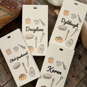 May include: Four rectangular, off-white kitchen signs with black text and illustrated baking icons. Each sign has a different name: "Douglene", "Dyldough", "Clint Yeaswood", and "Karen". A loaf of bread and other baking tools are in the background.