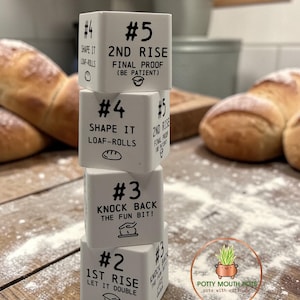 May include: A stack of four white ceramic cubes with baking instructions. Each cube has a number and text like "1st Rise" and "Knock Back". The cubes are on a wooden surface dusted with flour, with baked bread in the background.