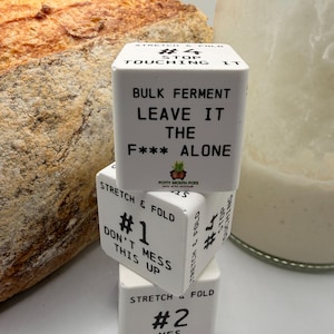 Könnte beinhalten: Drei gestapelte weiße Keramikwürfel, jeder mit schwarzem Text zum Thema Brotbacken. Der obere Würfel lautet: „#4 NICHT ANFASSEN MASSENFERMENTIEREN LASS ES DAS F*** ALLEIN“. Der untere Würfel sagt: „#2 JA WIEDER“. Ein Brotlaib ist im Hintergrund.