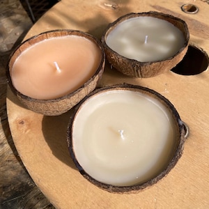 Könnte beinhalten: Drei Kerzen in Kokosnussschalen mit jeweils unterschiedlicher Wachsfarbe. Eine hat pfirsichfarbenes Wachs, eine andere cremefarbenes und die dritte hellbeige. Jede Kerze hat einen weißen Docht. Die Kerzen stehen auf einer Holzoberfläche.