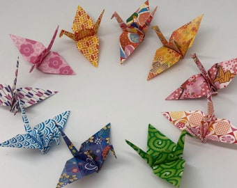 May include: A collection of ten origami paper cranes in various colors and patterns, including red, blue, green, yellow, and pink. The cranes are folded from traditional Japanese paper and are arranged in a circle on a white background.