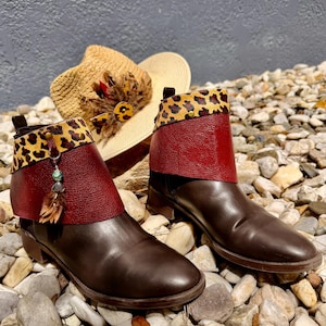 May include: A pair of ankle boots featuring dark brown leather lower sections, deep red textured leather cuffs, and leopard print trim. One boot includes a decorative charm with a turquoise bead and brown feathers, accompanied by a straw hat.