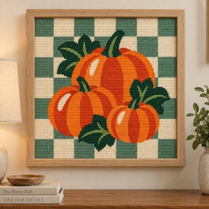 May include: A square needlepoint art piece in a light wood frame featuring three orange pumpkins with dark green leaves. The background consists of a cream and sage green checkered pattern. The needlepoint texture is visible throughout the design.