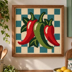Peut inclure: Une broderie au point de croix carrée encadrée en bois, représentant deux piments rouges et un piment vert central avec des feuilles vertes, sur un fond à motif damier de couleur crème clair et bleu sarcelle.