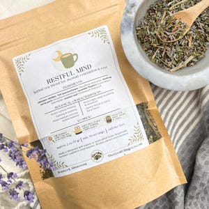 May include: A brown paper bag of "RESTFUL MIND" herbal tea blend for mental relaxation and rest. The bag is open, revealing the tea and text. A small bowl of dried herbs and a sprig of lavender are also visible.