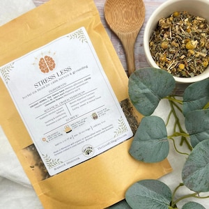 May include: A package of "Stress Less" herbal tea blend for calm nerves and grounding, with a label listing ingredients and brewing instructions. A wooden spoon and a bowl of loose tea are also visible, along with green leaves.