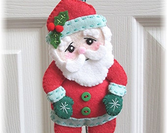May include: A felt Christmas ornament of Santa Claus wearing a red suit, white beard, and green mittens. He has a green holly sprig on his hat and two green buttons on his coat.
