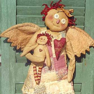 Puede incluir: Un ángel de trapo hecho a mano con piernas a rayas rojas y blancas y un vestido de retazos. El ángel sostiene una muñeca de trapo más pequeña con un corazón rojo en el pecho. La muñeca más grande tiene un parche en forma de corazón en su vestido que dice "Be My Mine".