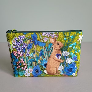 May include: A rectangular cosmetic bag with a vibrant floral and rabbit print. The bag features a brown rabbit holding a flower, surrounded by blue, purple, and white flowers on a green background. It has a green zipper and trim.