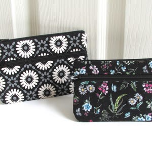 Può includere: Due pochette con cerniera. Una è nera con un motivo floreale bianco e grigio. L'altra è più piccola, nera e ricoperta da una colorata stampa floreale con fiori rosa, blu e bianchi e fogliame verde.