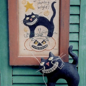 Può includere: Un ricamo incorniciato con un gatto nero e una zucca intagliata, con il testo "Halloween is.. Starry night, witches flight, jack-o-lanterns shining bright!" Un gatto nero di peluche è seduto su una zucca marrone con un viso di zucca intagliata.