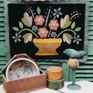May include: A black felt wall hanging with a colourful floral design in a yellow vase. The vase is on a black background with a scalloped edge. There are also three wooden boxes and a blue bird on a green stand.