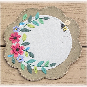 May include: A round felt coaster with a green leafy vine and pink and purple flowers around the edge. A bee with yellow and black stripes is flying above the flowers.