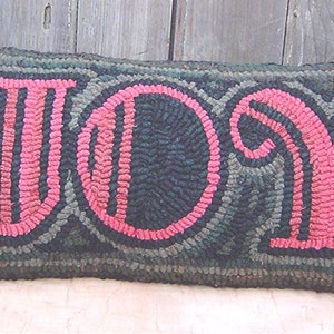 May include: A long, rectangular pillow with a dark green background and the word "JOY" in pink lettering. The letters are outlined in black and have a stylized, vintage font.