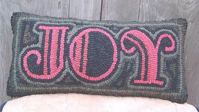 May include: A long, rectangular pillow with a dark green background and the word "JOY" in pink lettering. The letters are outlined in black and have a stylized, vintage font.