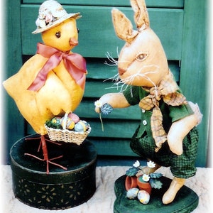 May include: Two Easter-themed fabric figures: a yellow chick wearing a hat and a red bow, and a tan bunny in green plaid clothing. The chick holds a basket of Easter eggs, and the bunny offers a flower. Both figures are on decorative bases.