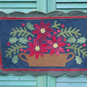 Może przedstawiać: Ręcznie robiona wisząca ozdoba ścienna z filcu z falistym brzegiem. Wzór przedstawia brązowy wazon wypełniony czerwonymi kwiatami poinsecji, zielonymi liśćmi ostrokrzewu i czerwonymi jagodami na ciemnoniebieskim tle.