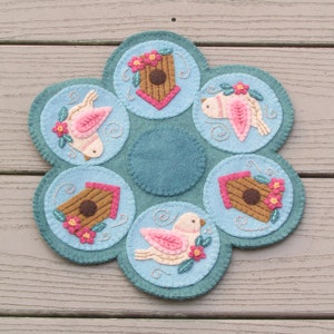 Può includere: Un arazzo da parete in feltro blu con un design circolare che presenta uccelli rosa, case degli uccelli marroni e fiori rosa. Il design è cucito con filo bianco.